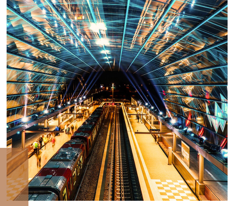 Blick in eine moderne Bahnhofshalle mit zwei Gleisen, einem roten Zug und einer gewölbten, beleuchteten Glasüberdachung in Blau- und Gelbtönen.