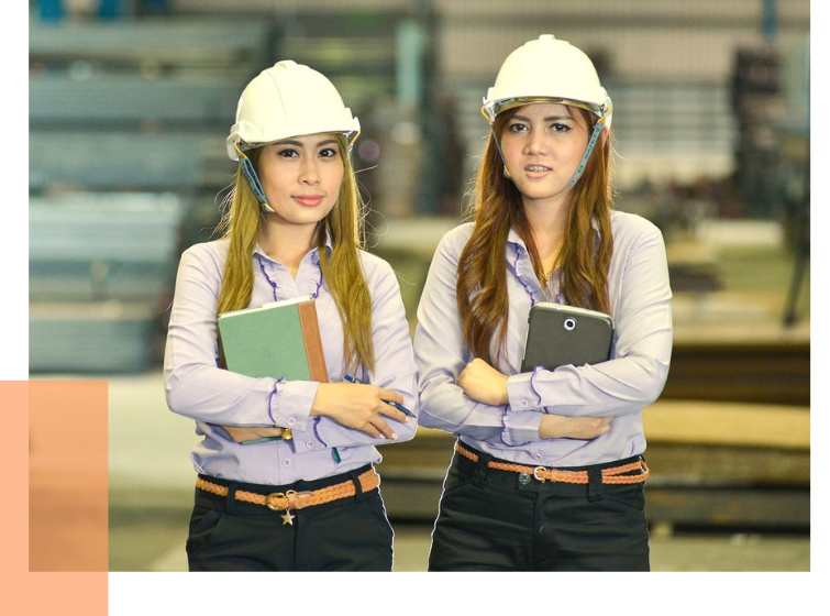 Zwei Frauen mit weißen Schutzhelmen, hellblauen Hemden und schwarzen Hosen stehen nebeneinander, eine hält ein Tablet, die andere einen Ordner.
