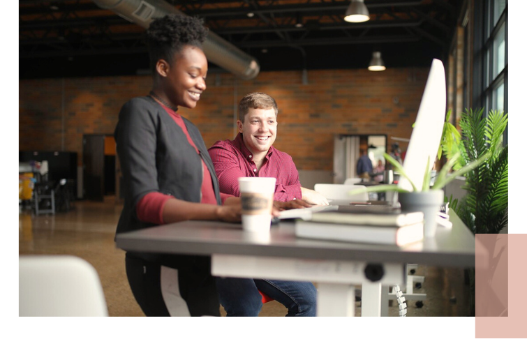 Zwei Personen sitzen an einem höhenverstellbaren Schreibtisch mit Laptop, Kaffeebecher und Pflanzen am Fenster in einem modernen Büro – ein Symbol für zugängliche, inklusive Arbeitsumgebungen im Sinne von UN-Ziel 10.