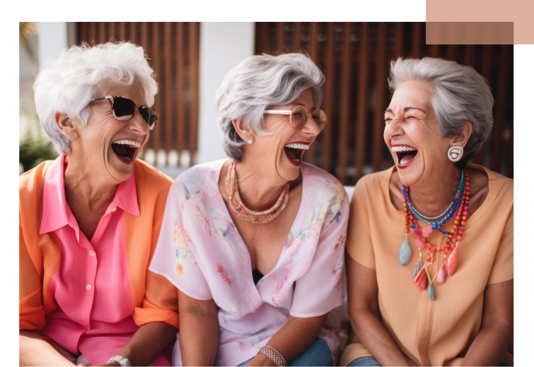 Drei ältere Frauen sitzen nebeneinander, tragen bunte Oberteile und auffälligen Schmuck, darunter Halsketten und Ohrringe.