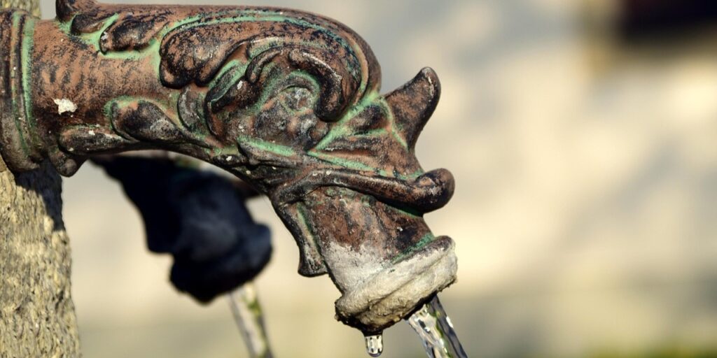 Nahaufnahme eines alten, dekorativen Metallwasserhahns in Form eines Drachenkopfs, aus dessen Maul Wasser fließt. Symbolisiert die Erhaltung traditioneller Wasserquellen und den kulturellen Wert des öffentlichen Zugangs zu sauberem Wasser.