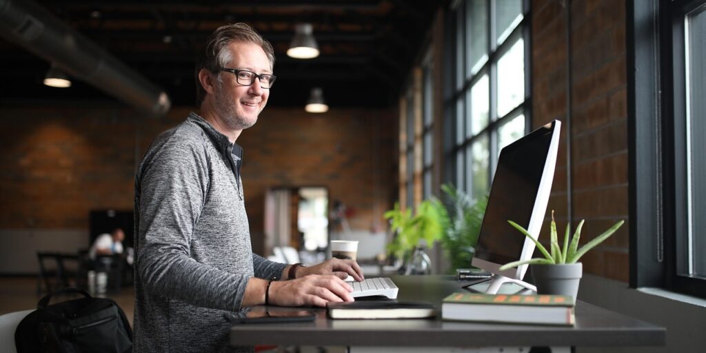 Person sitzt an Schreibtisch mit Tastatur, Monitor, Kaffeebecher, Notizbuch und Pflanze neben einem Fenster in einem Raum mit Backsteinwänden.