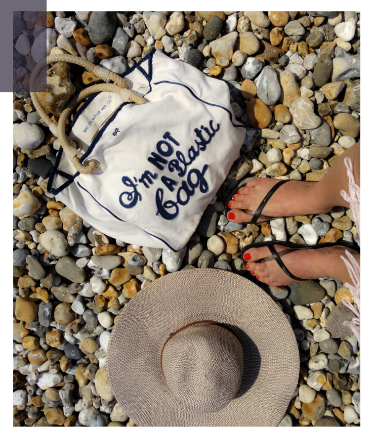 Stofftasche mit der Aufschrift ‚I’m not a plastic bag‘ liegt auf Kieselsteinen neben einem Sonnenhut und Füßen in Sandalen – Förderung von plastikfreien und wiederverwendbaren Alternativen. SDG 12