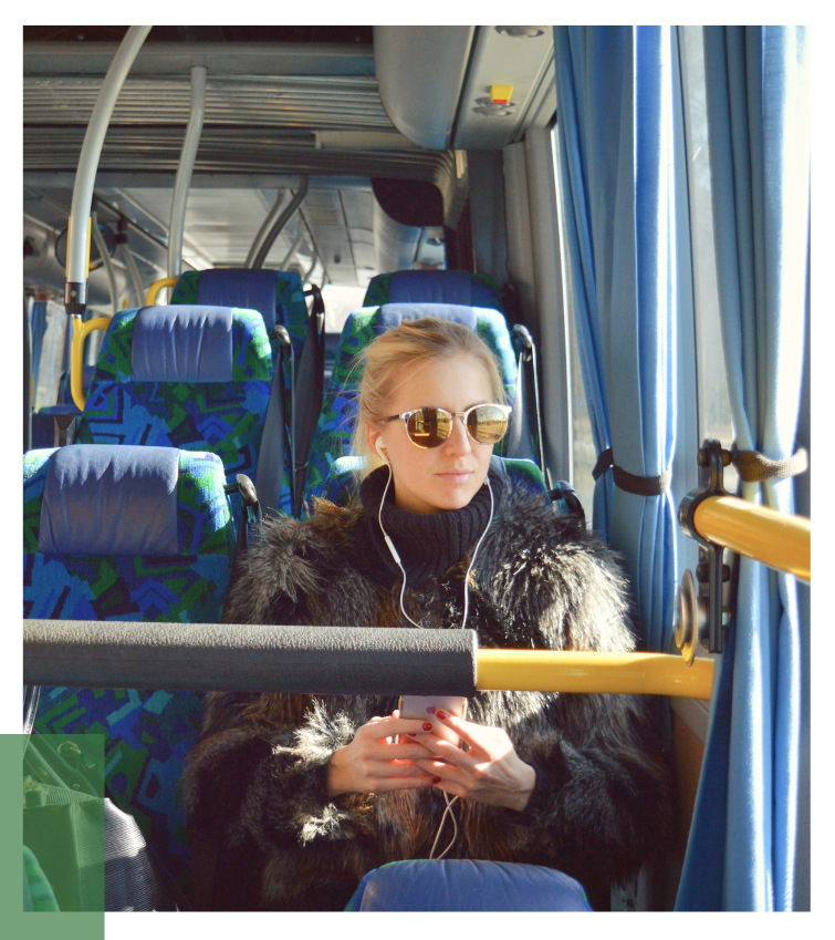 Person im schwarzen Mantel sitzt im Bus mit blau-grün gemusterten Sitzen und nutzt ein Smartphone – Darstellung klimafreundlicher Mobilität durch öffentliche Verkehrsmittel.