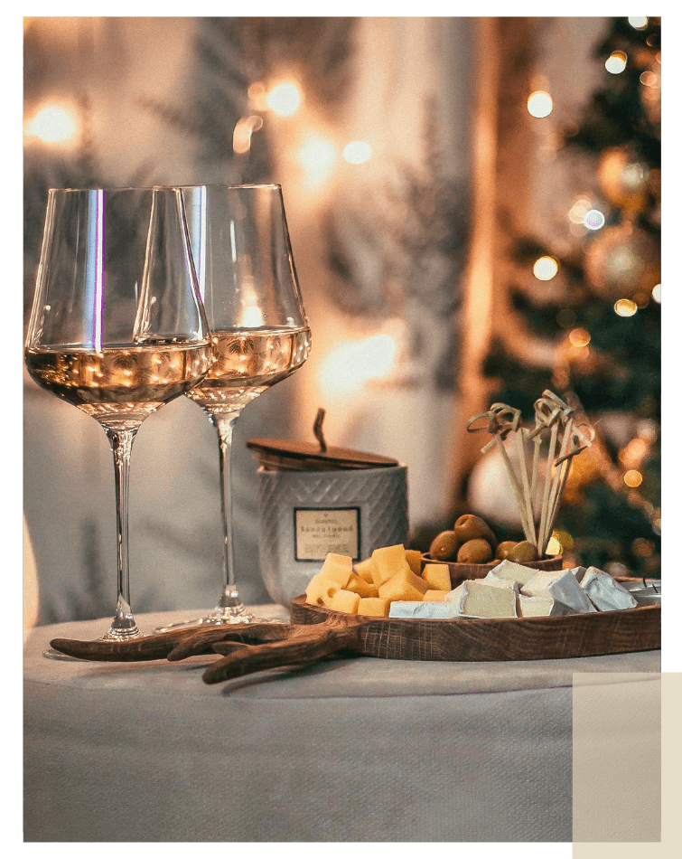 Zwei Weingläser mit Weißwein neben einer Holzplatte mit Käse und Oliven vor unscharfem Weihnachtsbaum und Lichterkette