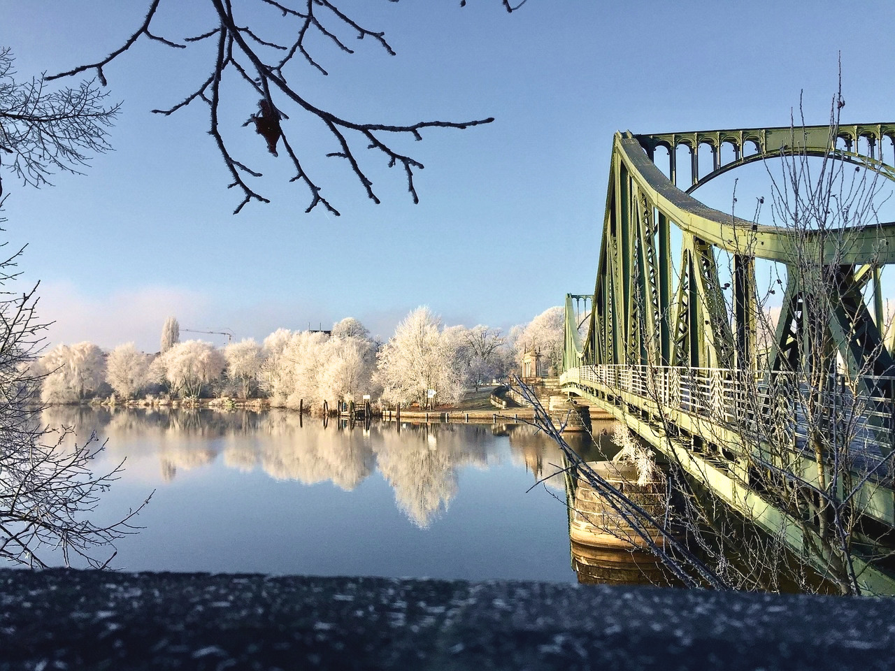 Grüne Stahlbrücke über ruhigen Fluss mit spiegelglatter Wasseroberfläche und schneebedeckten Bäumen im Winter