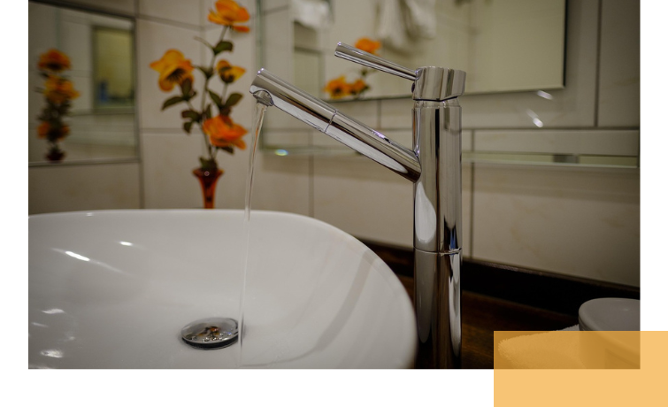 A modern, shiny stainless steel faucet with flowing water over a round sink. In the background, orange flowers are visible in a vase.