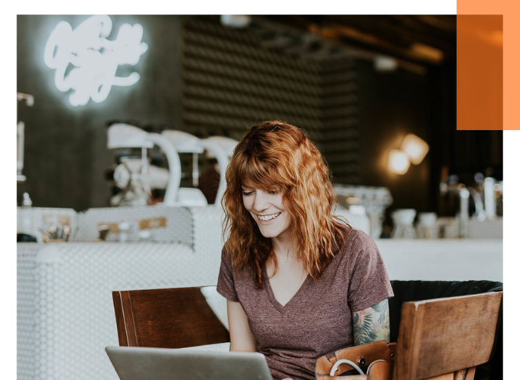 Person with curly hair and T-shirt sitting at a wooden table with laptop in a café or restaurant.