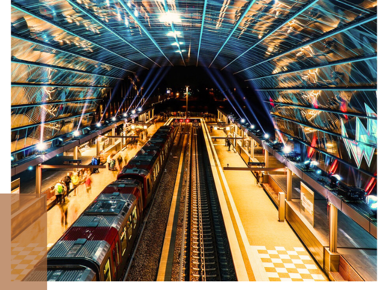 A view of a modern train station hall with two tracks, a red train, and a curved, illuminated glass canopy in shades of blue and yellow.