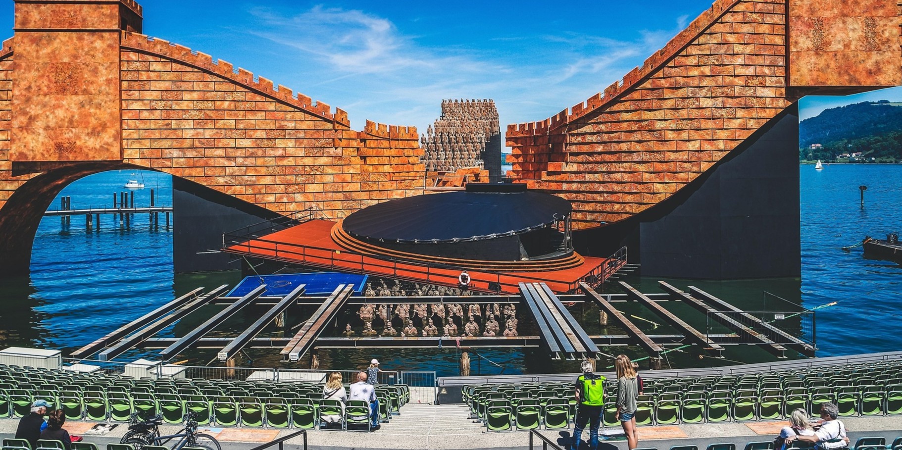 Stage setup with red brick walls and rows of chairs in front of water, people sitting and standing in the foreground.