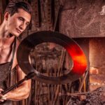 Muscular man wearing a black apron holding a large metal ring near a forge with bright orange flames.