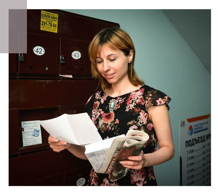 A woman in a floral dress stands in a dimly lit hallway holding a newspaper and a letter beside numbered mailboxes, reflecting everyday moments of visibility, and equal access.