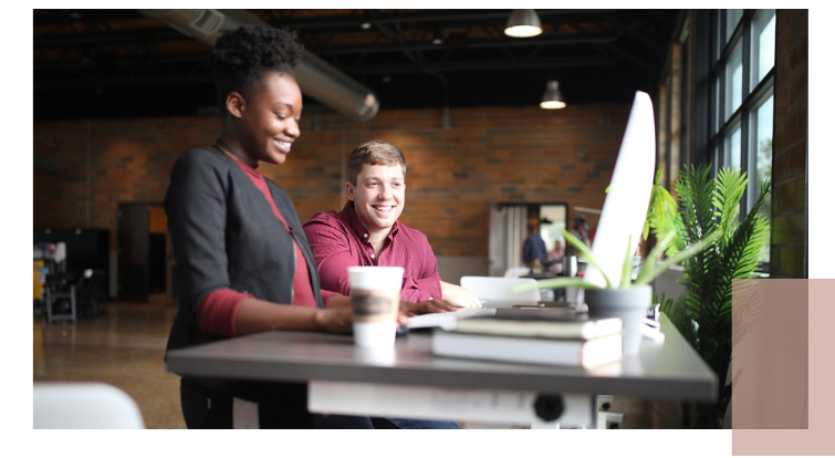 Two people sit together at a table with a laptop, coffee cup, and a small plant in a bright brick-walled room, reflecting an inclusive and welcoming setting.