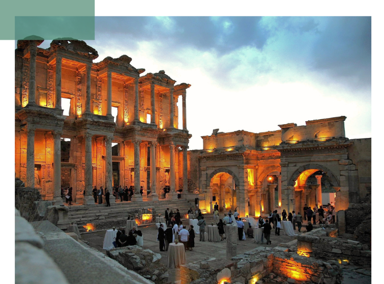 A historic, illuminated ancient ruins complex serves as the venue for an evening event with visitor groups. The image shows the preservation and use of cultural heritage as part of sustainable urban development.