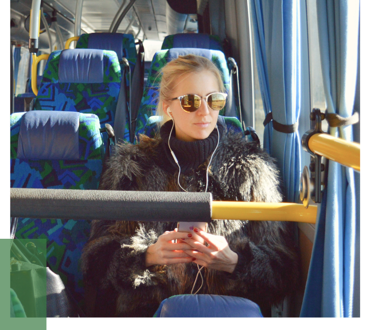 A person in a black coat sitting on a bus with blue-green patterned seats while using a smartphone, illustrating climate-friendly mobility through public transportation.