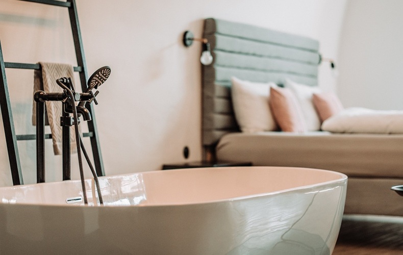 Freestanding white bathtub with black faucet in front of a bed with gray headboard and multiple pillows.